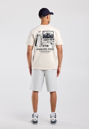 Man draagt een beige t-shirt met print van een vintage cassettebandje en grijze korte broek, van achteren gezien tegen een effen lichte achtergrond.