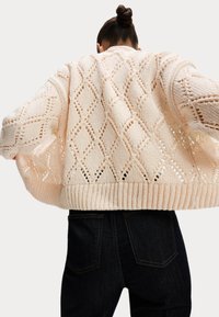 Häkelstrick-Jacke in hellem Pfirsich mit Rautenmustern, überschnittenen Schultern und geripptem Saum. Getragen über dunklen Jeans.
