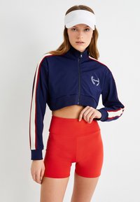 Chaqueta recortada azul marino con cremalleras y franjas blancas y rojas, que presenta un pequeño emblema. Combinada con pantalones cortos rojos de talle alto, que destacan por su textura suave.