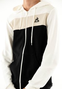 Felpa con zip dal design a blocchi di colore: bianco, crema e nero. Presenta una tasca a marsupio e il logo sul petto. Materiale in cotone.