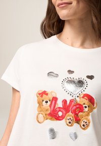 Weißes T-Shirt mit kurzen Ärmeln, das eine Grafik von zwei Teddybären, rotem "Liebe"-Text und metallischen Herzen zeigt. Weiches Material mit rundem Ausschnitt.