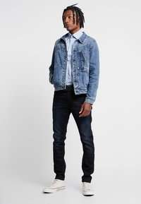 Hombre con rastas que lleva una camisa de botones azul claro, una chaqueta de mezclilla azul, jeans oscuros y zapatillas blancas, de pie contra un fondo liso.