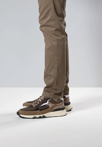 Ruskeat kuvioidut lenkkitossut, joissa on teksturoitu mesh ja sileät yksityiskohdat, paksulla pohjalla ja siksak-kuvioilla, käytettynä khakin väristen housujen kanssa.