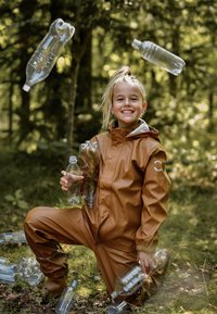 Hnědý nepromokavý pláštěnkový oblek s elastickými manžetami, držící plastové lahve, obklopený rozptýlenými lahvemi v lesním prostředí.