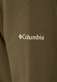 Olivgrön tyg med en slät textur har en vit "Columbia"-logotyp på sidan. Enkel, atletisk design utan ytterligare mönster.