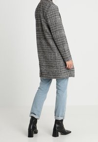 Person som bär en grå rutiga rock, ljusblå uppvikta jeans och svarta klackade ankelskor, stående mot en enfärgad vit bakgrund.