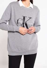 Grå sweatshirt med långa ärmar, med en stor svart "CK"-logotyp och texten "Calvin Klein Jeans". Bärs över en vit skjorta med krage.