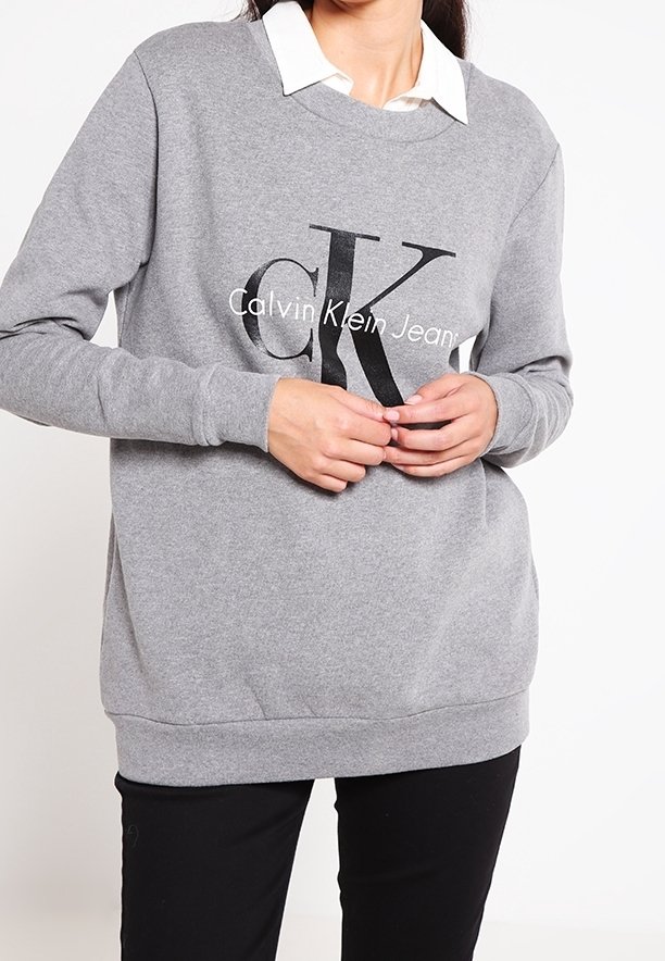Grå sweatshirt med långa ärmar, med en stor svart "CK"-logotyp och texten "Calvin Klein Jeans". Bärs över en vit skjorta med krage.