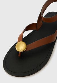 Sandali in pelle marrone con suola nera e un prominente accentuazione circolare dorata. Design piatto con fasce incrociate per una vestibilità sicura.