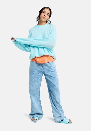 Svetlomodrý pletený nadrozmerný sveter s dlhými rukávmi, vrstvený cez oranžové tričko. Kombinovaný so svetlomodrými džínsami s širokými nohavicami a modrými sandálmi.