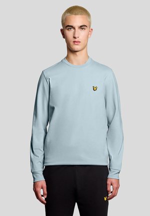 Ung mand med kort blondt hår iført en lyseblå langærmet skjorte og sorte bukser med små gule logoer, stående mod en ensfarvet baggrund.