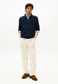 Mann in einem marineblauen Pullover mit Viertel-Reißverschluss über einem gestreiften Hemd, hellen Trousers und braunen Schuhen, der mit den Händen in den Taschen steht.