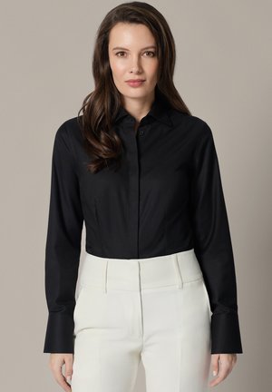 Kobieta z długimi brązowymi włosami, ubrana w czarną koszulę na guziki i białe spodnie z wysokim stanem na neutralnym, jednolitym tle.