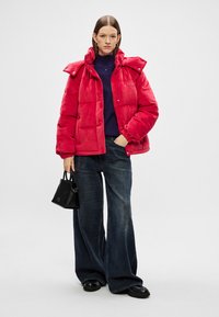 Sarkani pūkainis ar rāvējslēdzēju un pogu aizdari, ar plašu apkakli un elastīgām piedurknēm, apvienots ar platu tumsas džinsiem un melnu rokassomu.