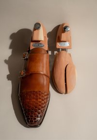 Braune Lederschuhe mit geflochtenem Zehendesign und doppeltem Schnallendetail, kombiniert mit einem Holzschuhbaum, der den Markennamen zeigt.