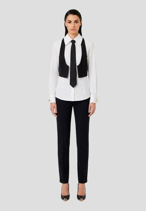 Donna in piedi che indossa una camicia bianca con cravatta nera e gilet nero senza maniche, pantaloni neri e scarpe nere con tacco alto e dettagli dorati.