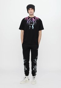 T-shirt nera con grafica di fulmine colorata, abbinata a pantaloni sportivi neri con design di fulmine simile. Entrambi sono realizzati in tessuto morbido.