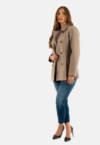 Cappotto peacoat beige in lana con design doppiopetto e bottoni in ottone, abbinato a jeans skinny blu e scarpe con tacco nere.