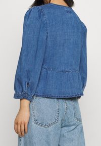 Modrý deninový peplum top s dlouhými nadýchanými rukávy, nařasenými detaily a kulatým výstřihem. Měkká textura a neformální design.