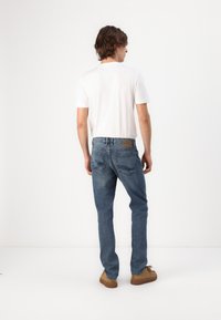 Jeans in denim blu con un design a gamba dritta, dotati di tasche posteriori e di una toppa in pelle beige in vita. Abbinati a una maglietta bianca.