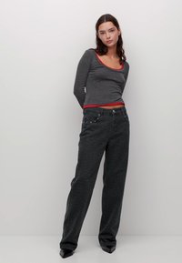 Giovane donna dai capelli scuri che indossa un top grigio a maniche lunghe con bordi rossi e jeans larghi a fantasia scura, in piedi contro un muro bianco.