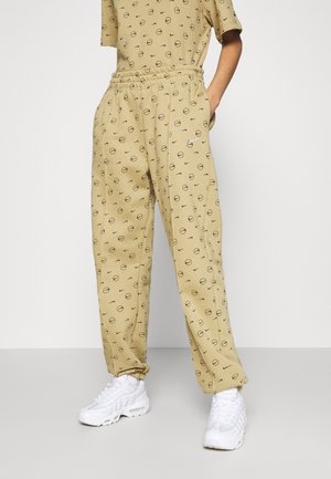 Personne portant un pantalon beige et un haut assorti avec des motifs circulaires noirs, baskets blanches, debout avec une main dans la poche.