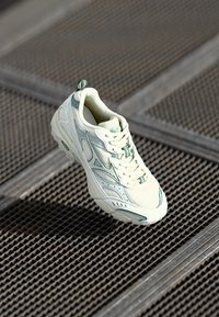 Zapatilla blanca con acentos verdes, material de malla, punta redondeada, suela texturizada y cordones. Presenta una lengüeta en la parte trasera y un diseño aerodinámico.