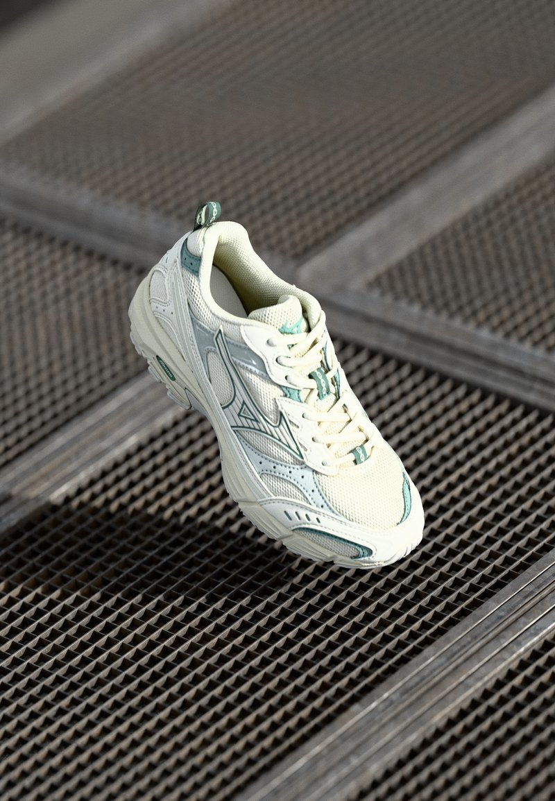 Zapatilla blanca con acentos verdes, material de malla, punta redondeada, suela texturizada y cordones. Presenta una lengüeta en la parte trasera y un diseño aerodinámico.