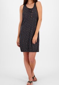 Kobieta w sukience bez rękawów o ciemnoszarym kolorze, sięgającej do kolan, z wielokolorowymi kropkami, trzema guzikami na klatce piersiowej i czarnymi sandałami.