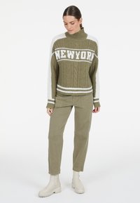 Olivgrüner Strickpullover mit weißen Akzenten und dem Text "NEW YORK". Kombiniert mit olivgrünen Hosen und beigen Stiefeletten.