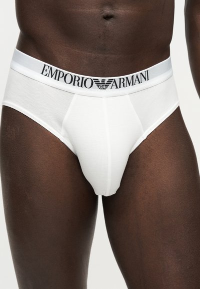 Αρσενικός κορμός που φοράει λευκά εσώρουχα Emporio Armani με λαστιχένια μέση με λογότυπο σε απλό φόντο.