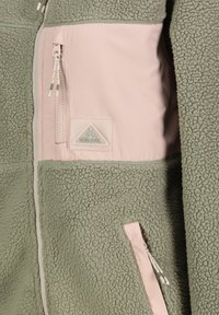 Olive green fleeced jacket with pink nylon accents, featuring two zippered pockets and a logo patch. Textured fabric with a soft finish.
