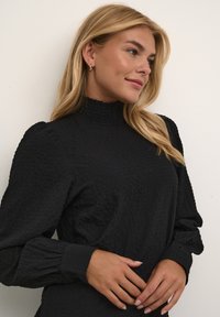 Haut noir texturé avec un col haut et des manches bouffantes, présentant des détails froncés au niveau de l'encolure et des poignets. Tissu lisse avec un motif subtil.