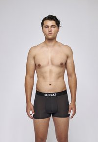 Junger Mann mit nassem Haar steht fröhlich blickend, oberkörperfrei, in schwarzen Snocks Boxer-Shorts vor einem schlichten grauen Hintergrund.