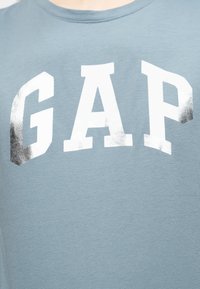 Ανοιχτό μπλε ύφασμα με μεγάλα λευκά και ασημί αντανακλαστικά γράμματα που σχηματίζουν τη λέξη "GAP" στην περιοχή του στήθους ενός πουκαμίσου.