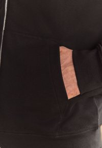 Melns zip-up kapucenieks ar priekšējo kabatu, kurā ir ievietota roka, izgatavots no mīkstas auduma ar gludu tekstūru un minimālu dizainu.