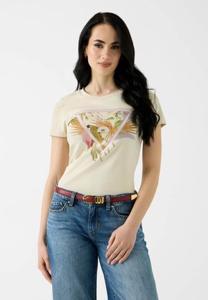 Frau mit langen schwarzen Haaren, die ein beiges T-Shirt mit floralem Muster, blaue Jeans und einen roten Gürtel vor einem einfarbigen hellen Hintergrund trägt.
