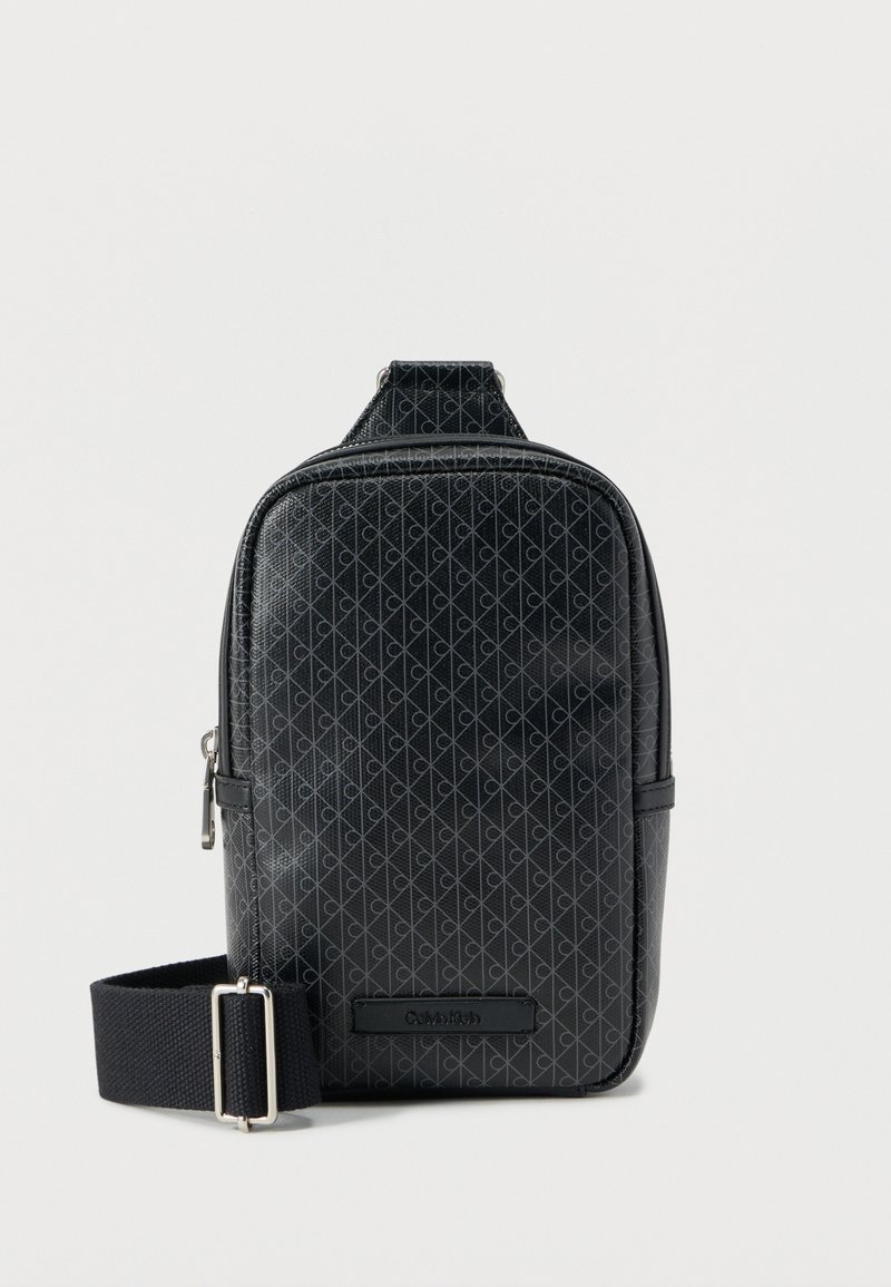 Μαύρη συνθετική τσάντα ώμου με σχέδιο διαμαντιού, κλείσιμο με φερμουάρ και ρυθμιζόμενο λουρί. Διακριτικό λογότυπο μπροστά. Λείο υλικό.