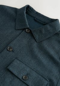 Camicia in morbida lana di un verde scuro, dotata di collare, due bottoni neri e una tasca sul petto. Texture liscia, design minimalista e senza motivi visibili.