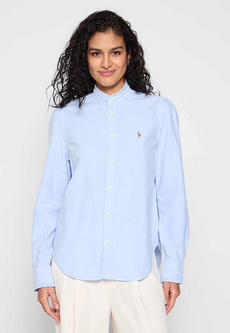 Frau mit lockigem Haar, die eine hellblaue gebuttonte Bluse und cremefarbene Hosen trägt, steht vor einem schlichten weißen Hintergrund.