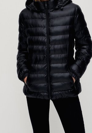 Black quilted hooded puffer jacket with front zipper and side pockets, worn by a person with hands in pockets against a plain background.
