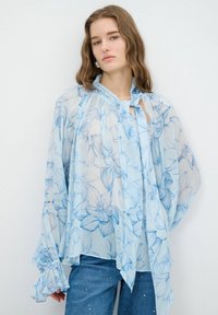 Lysblå, gennemsigtig bluse med blomsterprint, flæsekantede ærmer og snorekrave. Kombineret med denimjeans med små udsmykninger.