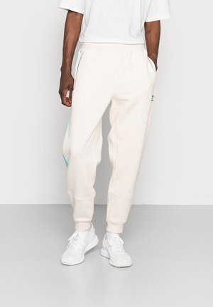 Mężczyzna w jasnobeżowych spodniach dresowych z bocznymi paskami i białych trampkach, stojący na jednolitym jasnym tle.