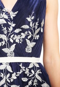 Námořnické modré šaty s jemným bílým květinovým vyšíváním. Měkčí látka s plisovanou texturou a bílým pásem, který zvýrazňuje pas.