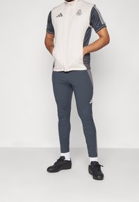 Chándal deportivo gris que presenta un chaleco de color claro con cremallera, tres franjas en las mangas y un logo. Unos zapatos deportivos negros completan el look.