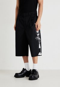 O persoană poartă pantaloni scurți largi negri cu un grafic alb pe lateral, pantofi negri cu studs, șosete albe și un top negru.