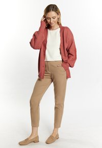 Cardigan lavorato a maglia color ruggine con un motivo testurizzato, chiusura a bottoni, indossato sopra una maglietta bianca e pantaloni beige sartoriali, abbinato a ballerine marrone chiaro.