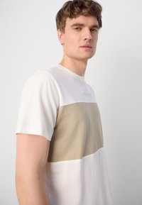 Lühikese varrukaga t-särk valge ja beeži värviga, värvibloki disainiga, millel on tekstuuriga kangas ja reljeefne logo ülemises osas.