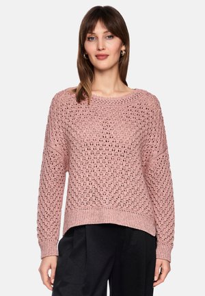 Frau trägt einen rosa offenen Strickpullover und schwarze Hose, steht vor einem schlichten hellen Hintergrund mit neutralem Gesichtsausdruck.