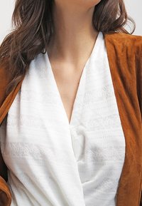 Bruine suède jas over een witte textuurtop met een V-hals. De top heeft subtiele patronen en fijne details door de stof heen.